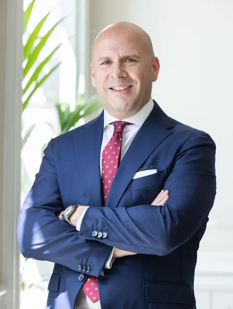 Mr Marco Scarci, thoracic surgeon, in a navy suit with a red polka‑dot tie standing indoors with arms crossed